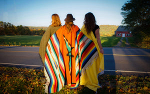 Three models shown from behind, each with handwoven Mexican blanket draped around their shoulders. Blanket on center model has Thunderbird design in deep orange with brightly colored stripes. Blanket on left model is solid Olive and right is solid Yellow.