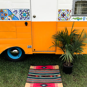 Door and side of vintage trailer, painted bright orange and white with hand painted Talavera tile decorations. Brightly colored handwoven rug and potted palm next to door.