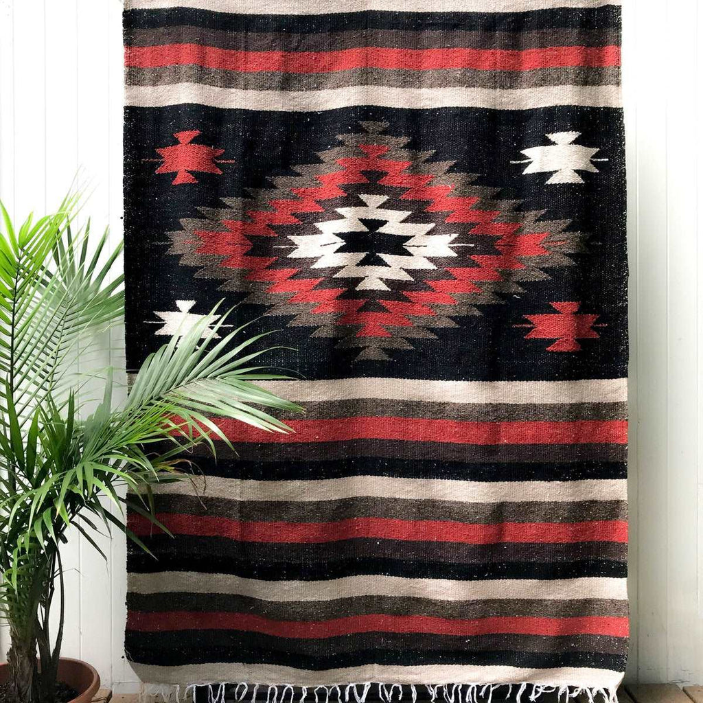 Mexican blanket woven in geometric design with center diamond and stripes in black, rust, brown, and tan with white fringe shown hanging against white wall with palm plant at side