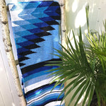 Mexican blanket handwoven in blue, black and white diamond and stripe pattern, hanging over birch ladder next to palm plant