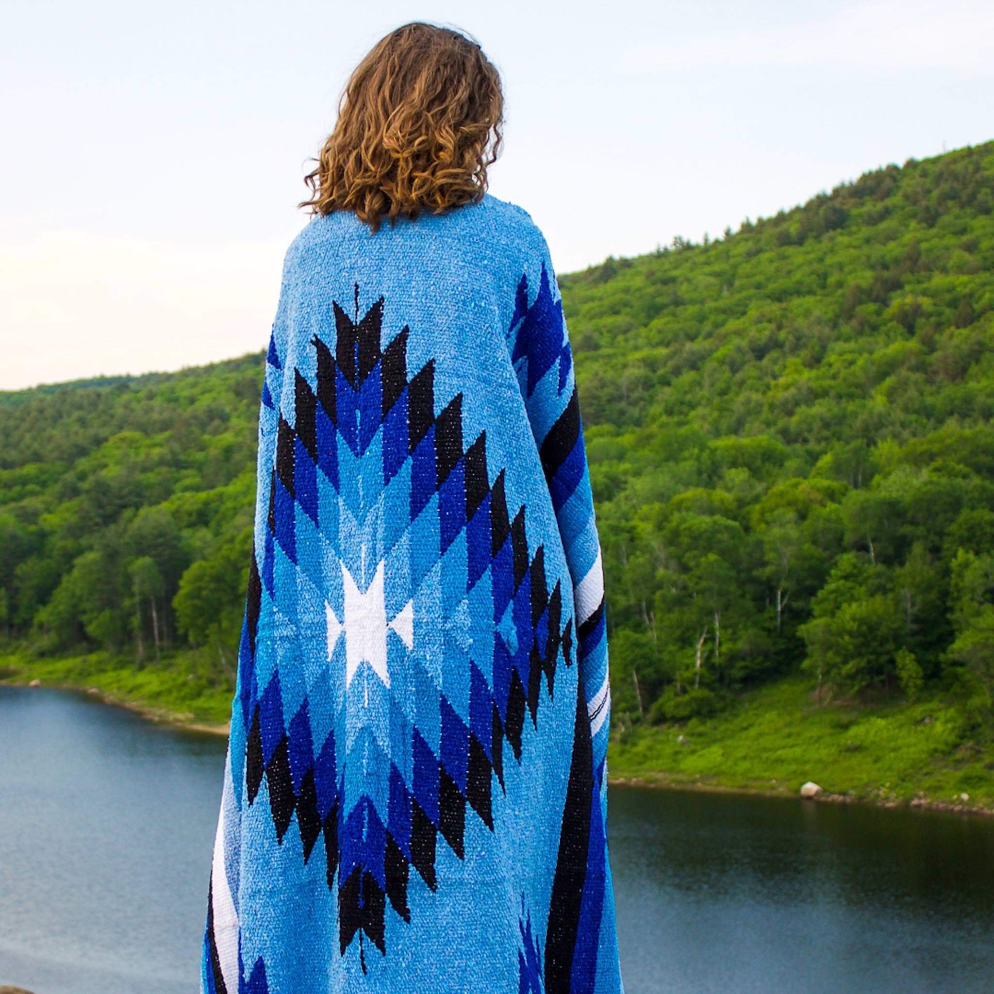 Model shown from behind wrapped in handwoven Mexican blanket with diamond and stripe design in shades of blue, black and white with mountain and lake background