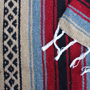 Close-up of blanket woven with alternating solid stripes in black, grey, burgundy and tan, and tan stripe with pattern of small black diamonds, edge of blanket has white fringe.
