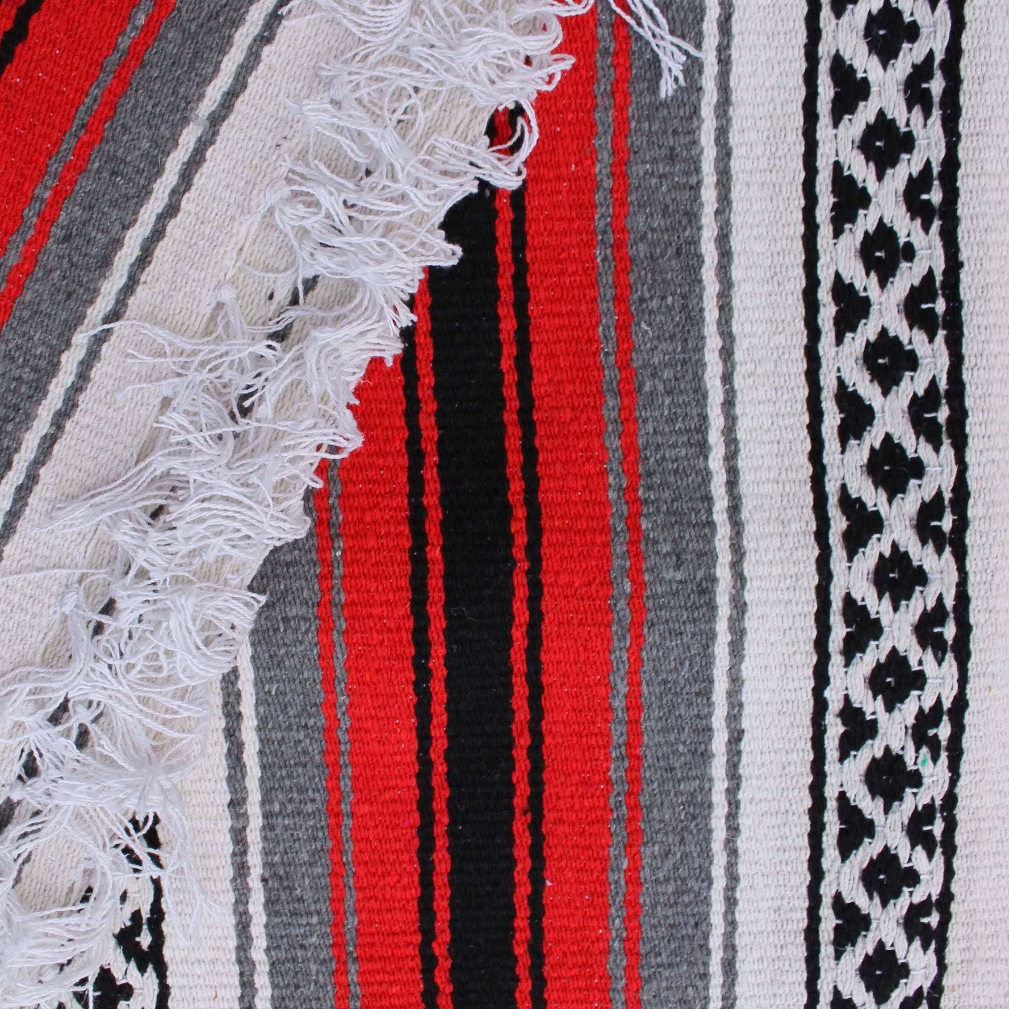Close-up of blanket woven with alternating solid stripes in white, black, grey and chocolate brown, and white stripe with pattern of small black diamonds, edge of blanket has white fringe.