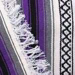 Close-up of blanket woven with alternating solid stripes in white, black, grey and bright purple, and white stripe with pattern of small black diamonds, edge of blanket has white fringe.