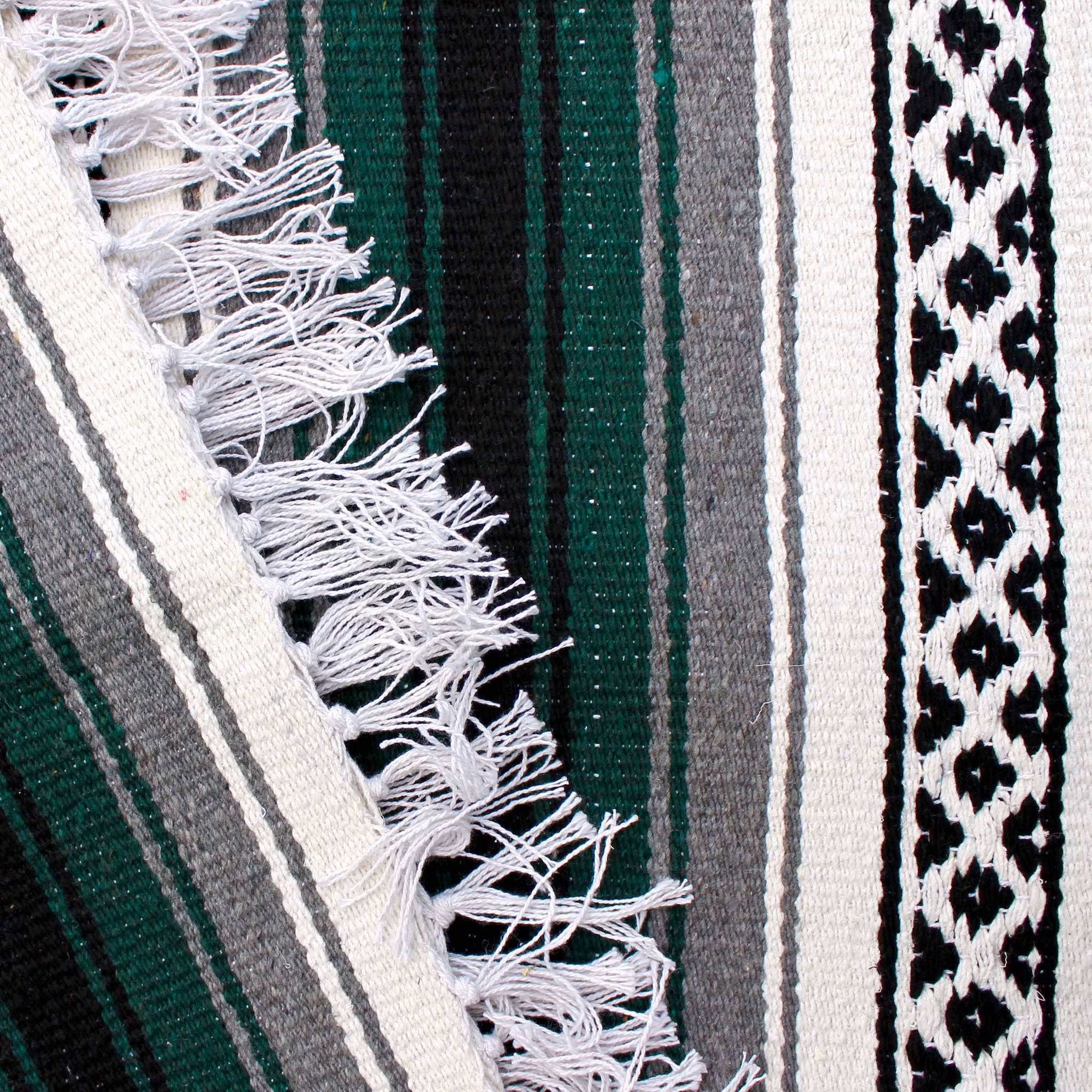 Close-up of blanket woven with alternating solid stripes in white, black, grey and hunter green, and white stripe with pattern of small black diamonds, edge of blanket has white fringe.