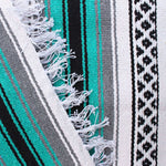 Close-up of blanket woven with alternating solid stripes in white, black, grey and bright teal, and white stripe with pattern of small black diamonds, edge of blanket has white fringe.