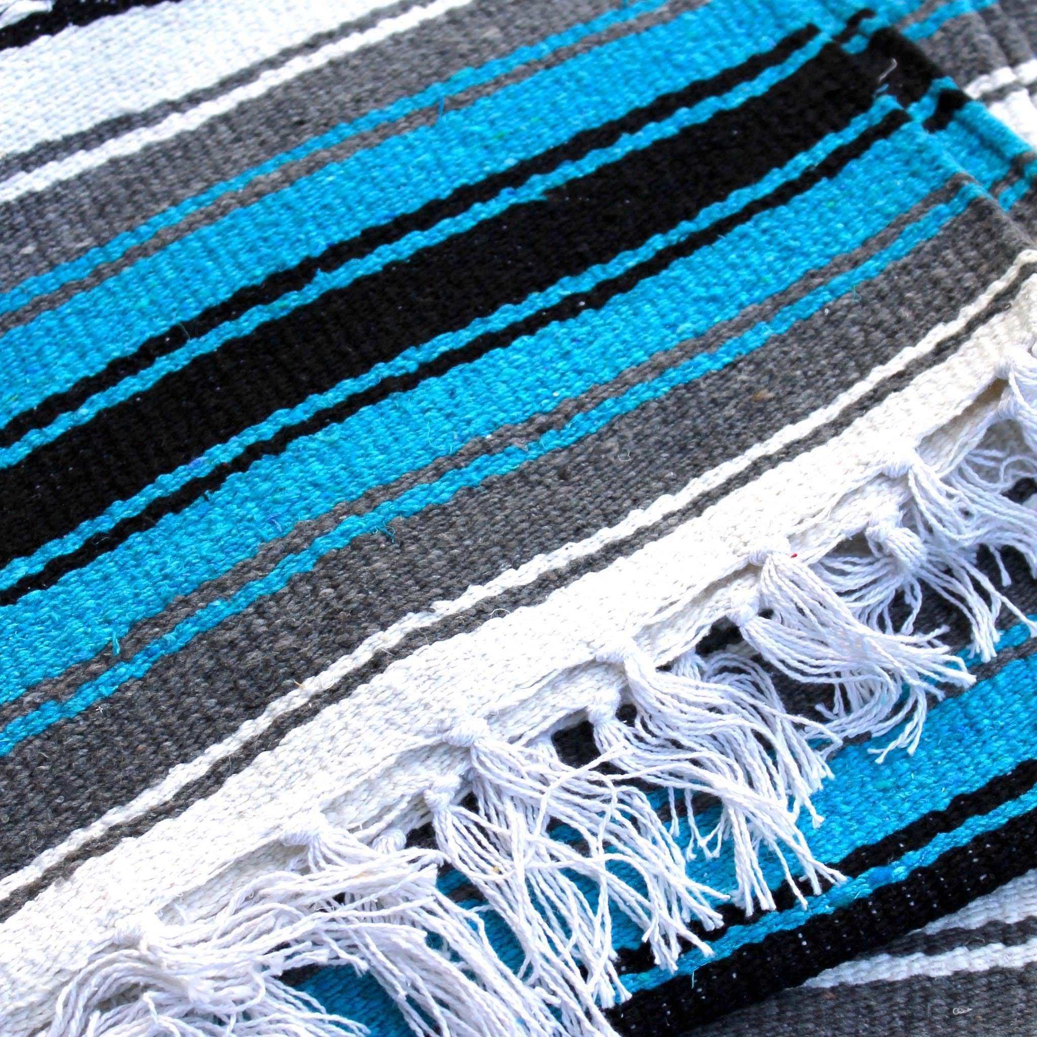 Close-up of blanket woven with alternating solid stripes in white, black, grey and bright turquoise, and white stripe with pattern of small black diamonds, edge of blanket has white fringe.