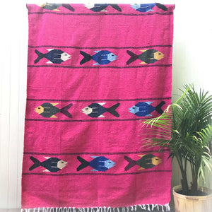 Woven blanket in hot pink with pattern of three multicolored fish in four alternating stripes, with white fringed edge, shown hanging against white wall with potted palm plant at side.
