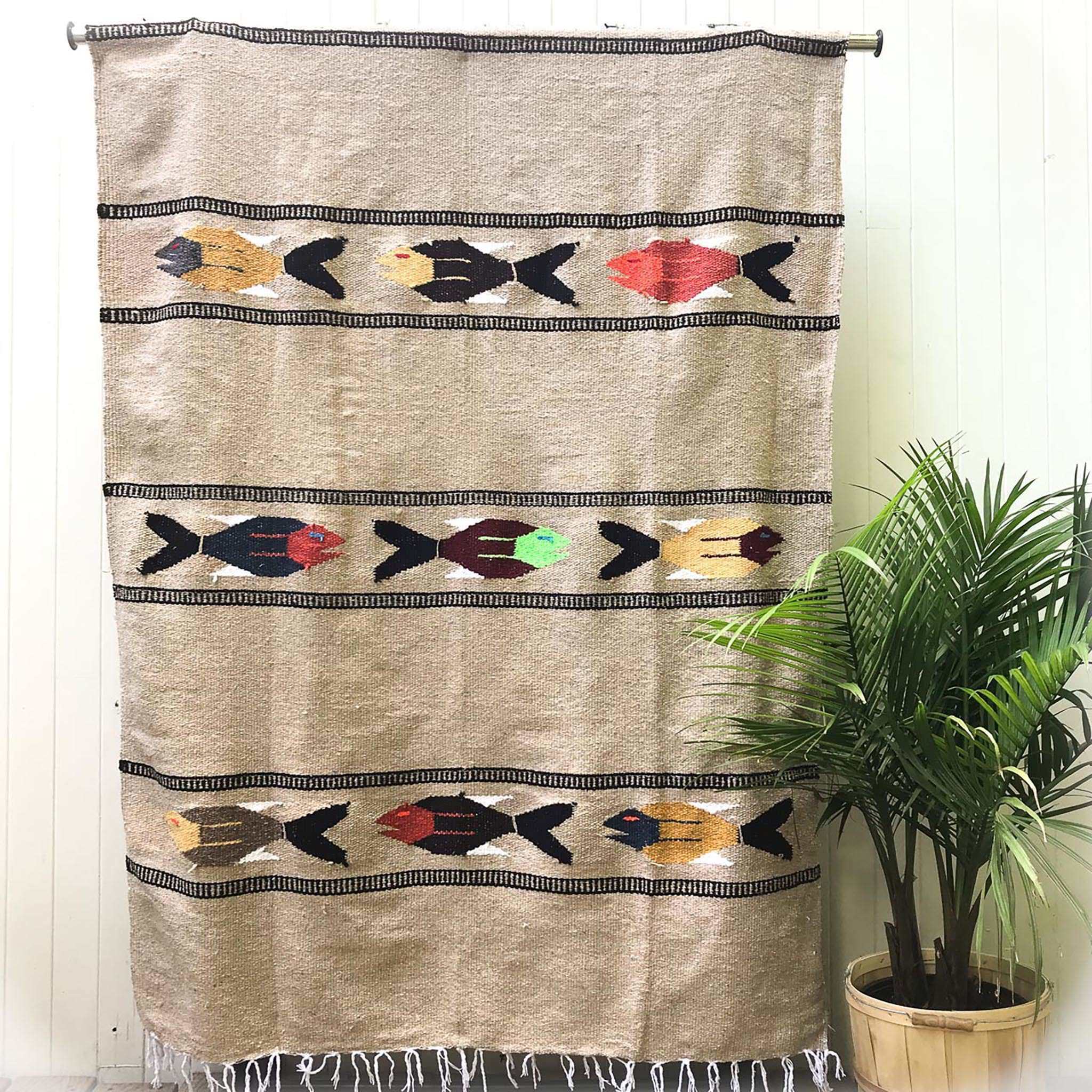 Woven blanket in natural tan with pattern of three multicolored fish in four alternating stripes, with white fringed edge, shown hanging against white wall with potted palm plant at side.