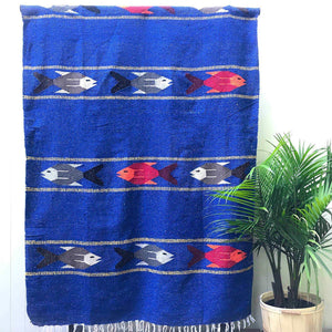 Woven blanket in royal blue with pattern of multicolored fish in four stripes, alternating with wide stripes of royal blue, with white fringed edge, shown hanging against white wall with potted palm plant at side.