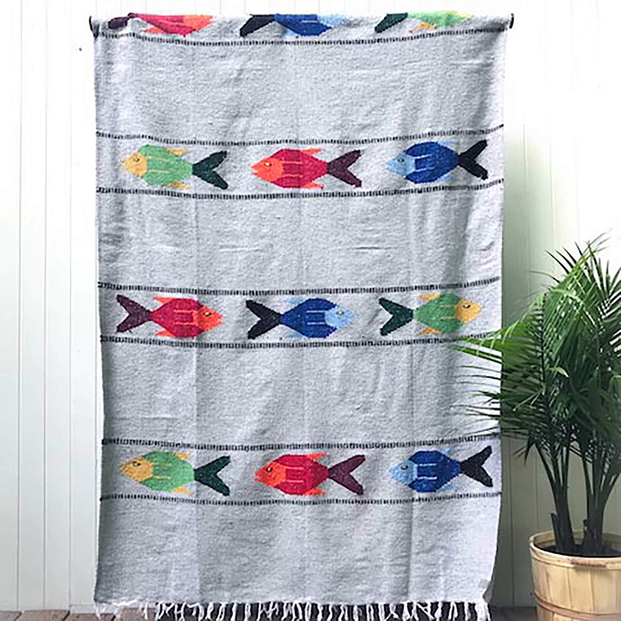 Woven blanket in light grey with pattern of three multicolored fish in four alternating stripes, with white fringed edge, shown hanging against white wall with potted palm plant at side.