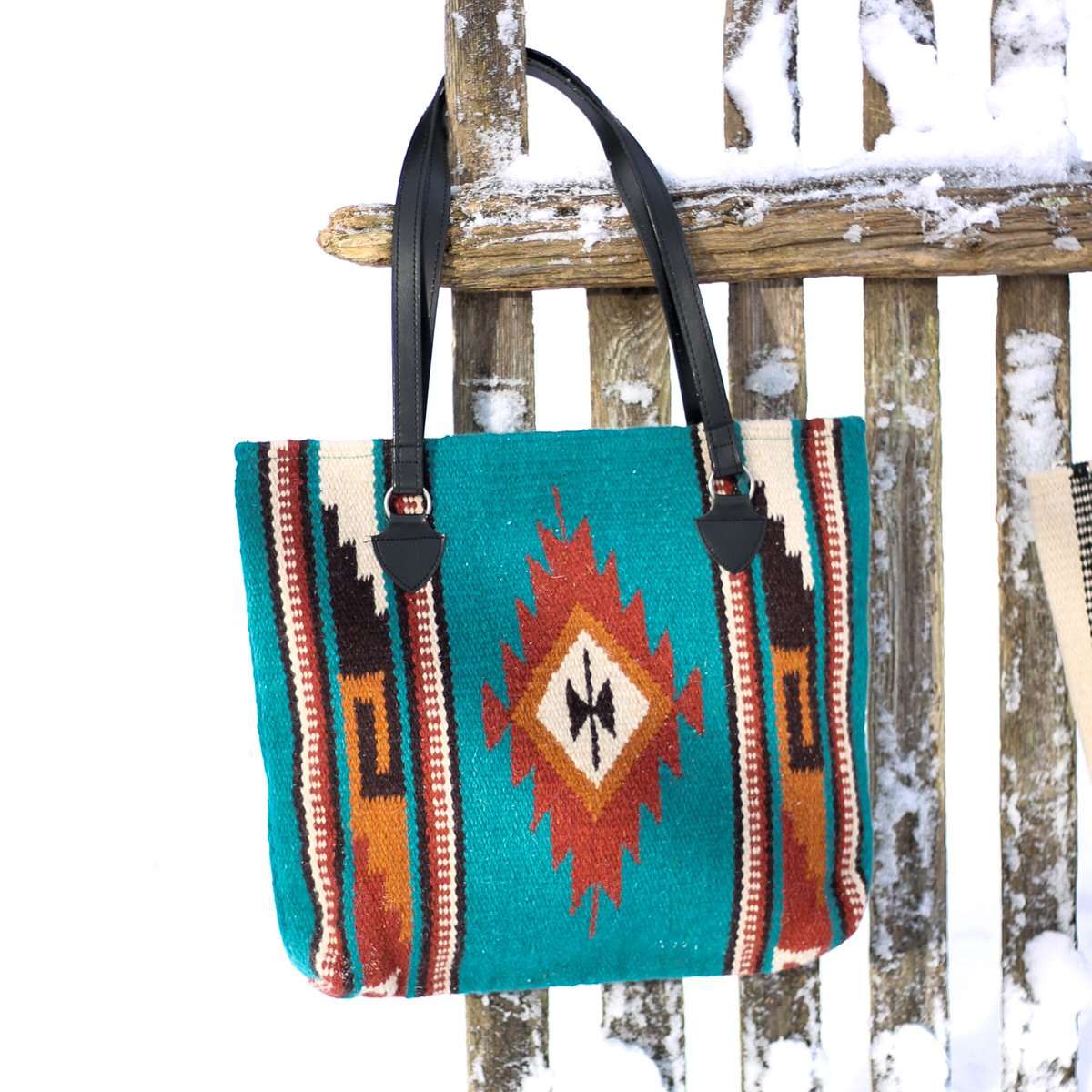 Handwoven wool handbag with center diamond design in turquoise and rust with black, cream and tan accents and black straps hanging from snow covered wood fence.