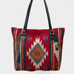 Handwoven wool handbag with center diamond design in burnt orange, blue and cream on deep red background, with geometric stripe design on each side and brown strap handle against white background.