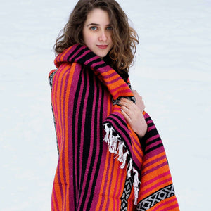 Young woman with brown hair, blue eyes, pale skin shown from waist up, looking into camera, hand on chest holding dark pink, orange, black striped blanket with white fringe draped around her shoulders