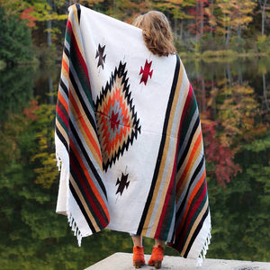 Woman with light brown hair shown from behind with San Miguel blanket draped around her shoulders, one arm extended up holding blanket, showing blanket with stripes and center diamond design in colors of rust, tan, black, dark green, grey, burgundy and white, standing against background of autumn leaves reflected in water.