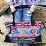 Woman shown from front, waist to knee, dressed in purplish grey sweater and ripped blue jeans, with hands holding straps of large rectangular weekender bag made of woven material, primarily grey with stripe and diamond design in dark green, cobalt blue, tan, orange, red, turquoise and black, with black suede handles and bottom corners, standing against background of brown rock wall.