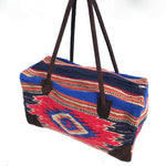 Rectangular weekender bag made of woven material, primarily red with stripe and diamond design in cobalt blue, dark orange, burgundy, tan and black, with brown suede handles and bottom corners against white background.