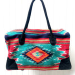 Rectangular weekender bag made of woven material, primarily teal and red with diamond and stripe design in orange, tan, turquoise, navy blue, white and black, with black suede handles and bottom corners against white background.