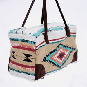 Rectangular weekender bag made of woven material, primarily tan and white with diamond and stripe design in orange, dark red, turquoise and black, with brown suede handles and bottom corners against white background.