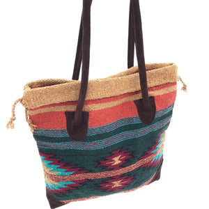 Square tote bag made from woven material, primarily dark green with diamond design in burgundy, turquoise, gold and black, with accent stripes in the same colors, handles and bottom corners in dark brown suede, shown against white background.