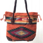 Square tote bag made from woven material in primarily dark orange with diamond design in gold, grey, burgundy, dark green and black, with accent stripes in the same colors, handles and bottom corners in dark brown suede, shown against white background.