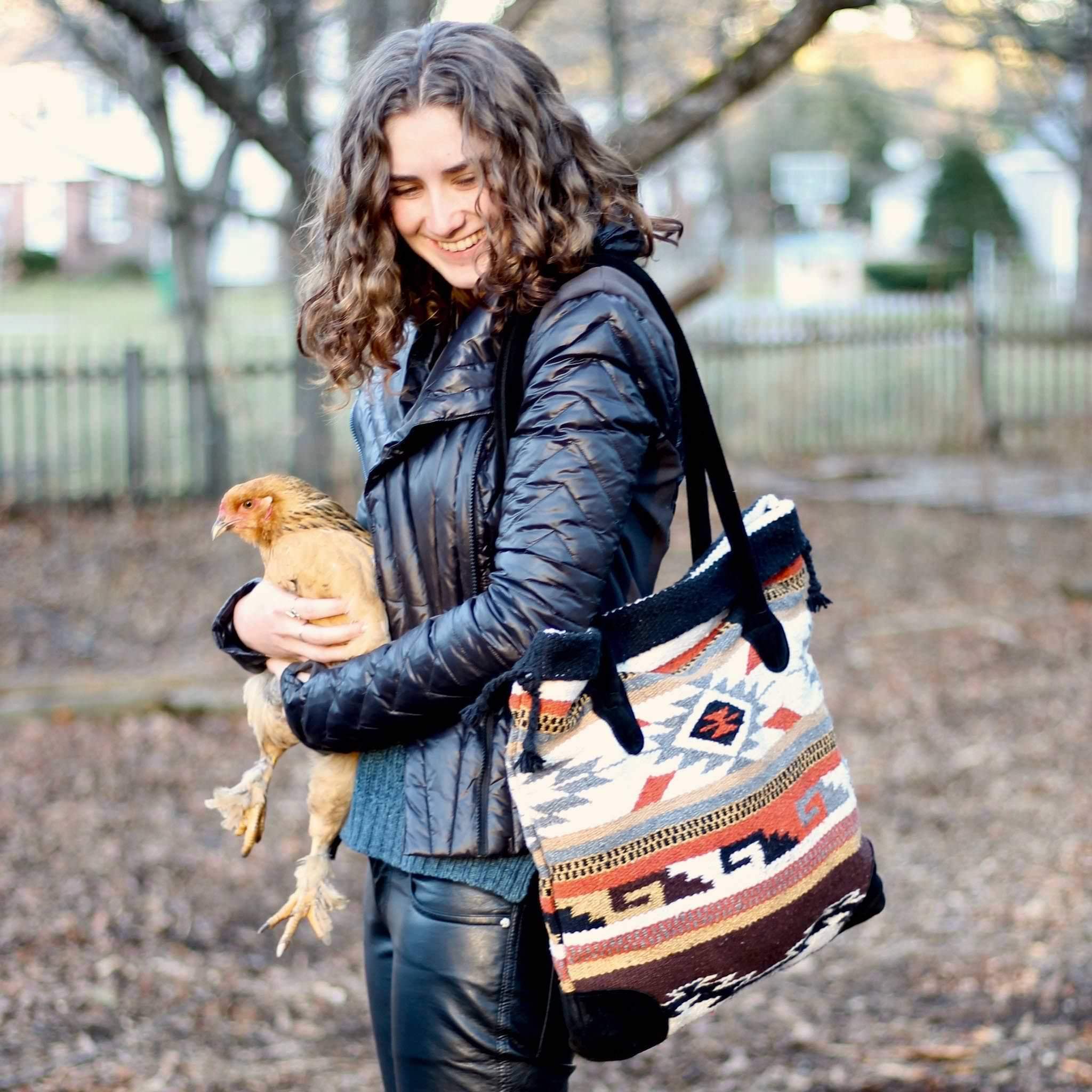 Smiling young woman with light brown wavy hair dressed in black leather jacket and pants, holding an orange chicken and wearing a square shoulder bag with geometric design in dark orange, grey, black, brown and white with black suede straps and bottom corners, against blurred background of bare trees and brown ground.