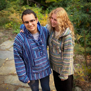 Young male and female couple standing side by side with arms around each other, wearing Baja hoodie jackets and dark pants. His hoodie is woven in narrow stripes of dark blue, light blue and white, and hers is cream, olive, turquoise and dark blue. Green foliage in background.