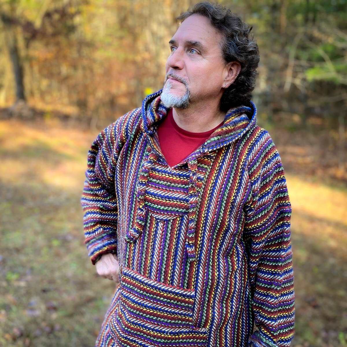 Male model with curly dark brown hair and grey goatee, wearing a Baja hoodie jacket with narrow stripes, woven in shades of red, purple, dark blue, yellow and white. Grass and trees in background.