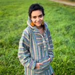 Female model with short dark hair wearing Baja hoodie jacket, striped in shades of teal, turquoise, dark blue, and cream. Green grass in background