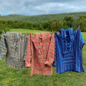 Three Baja hoodies,  hanging on clothesline with meadow and green hills in background. Hoodies are woven with a narrow striped pattern, one in shades of green, one in pinks and browns, and one in shades of blue.